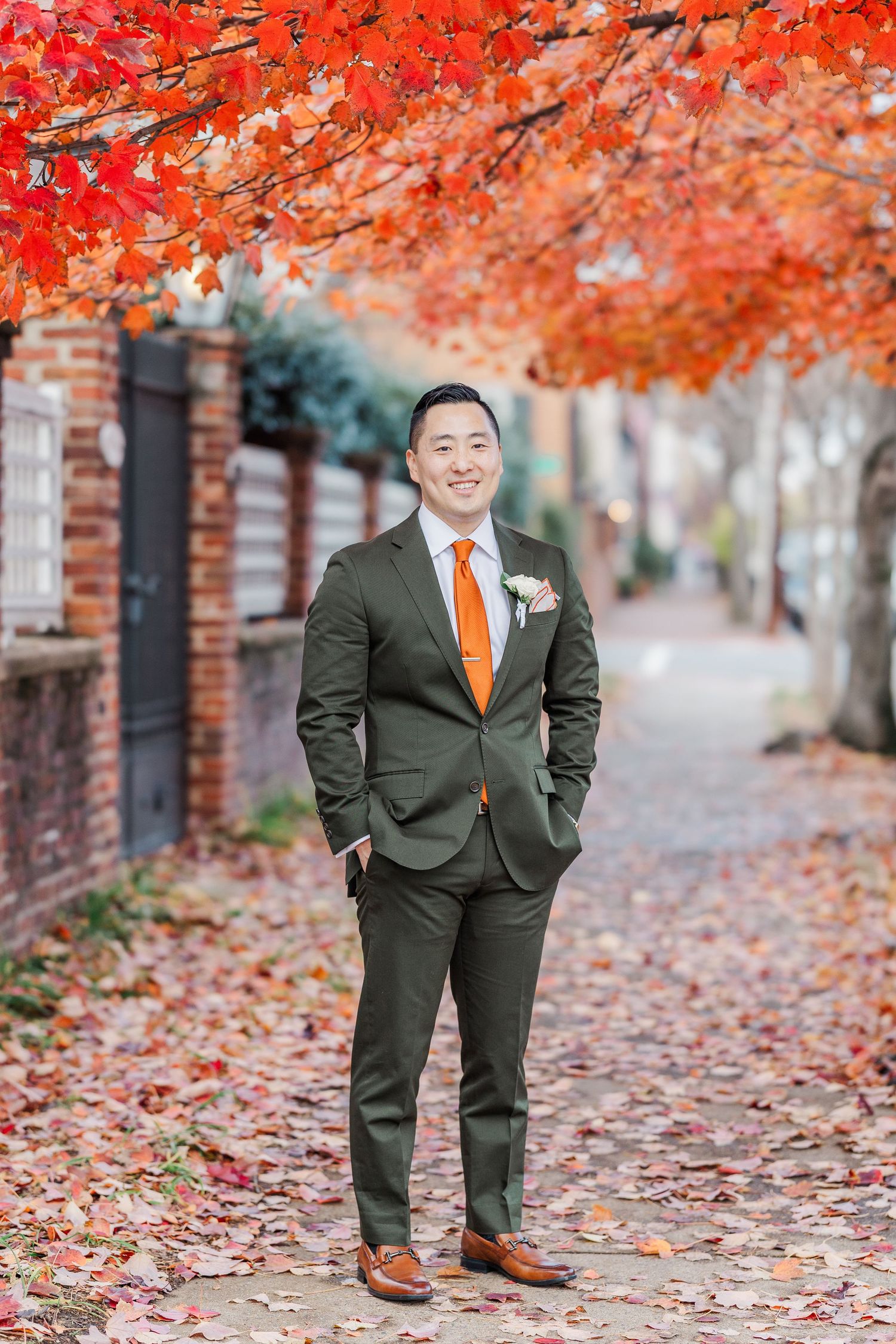 A person in a dark suit and orange tie stands on a leaf-covered sidewalk beneath vibrant red autumn foliage.