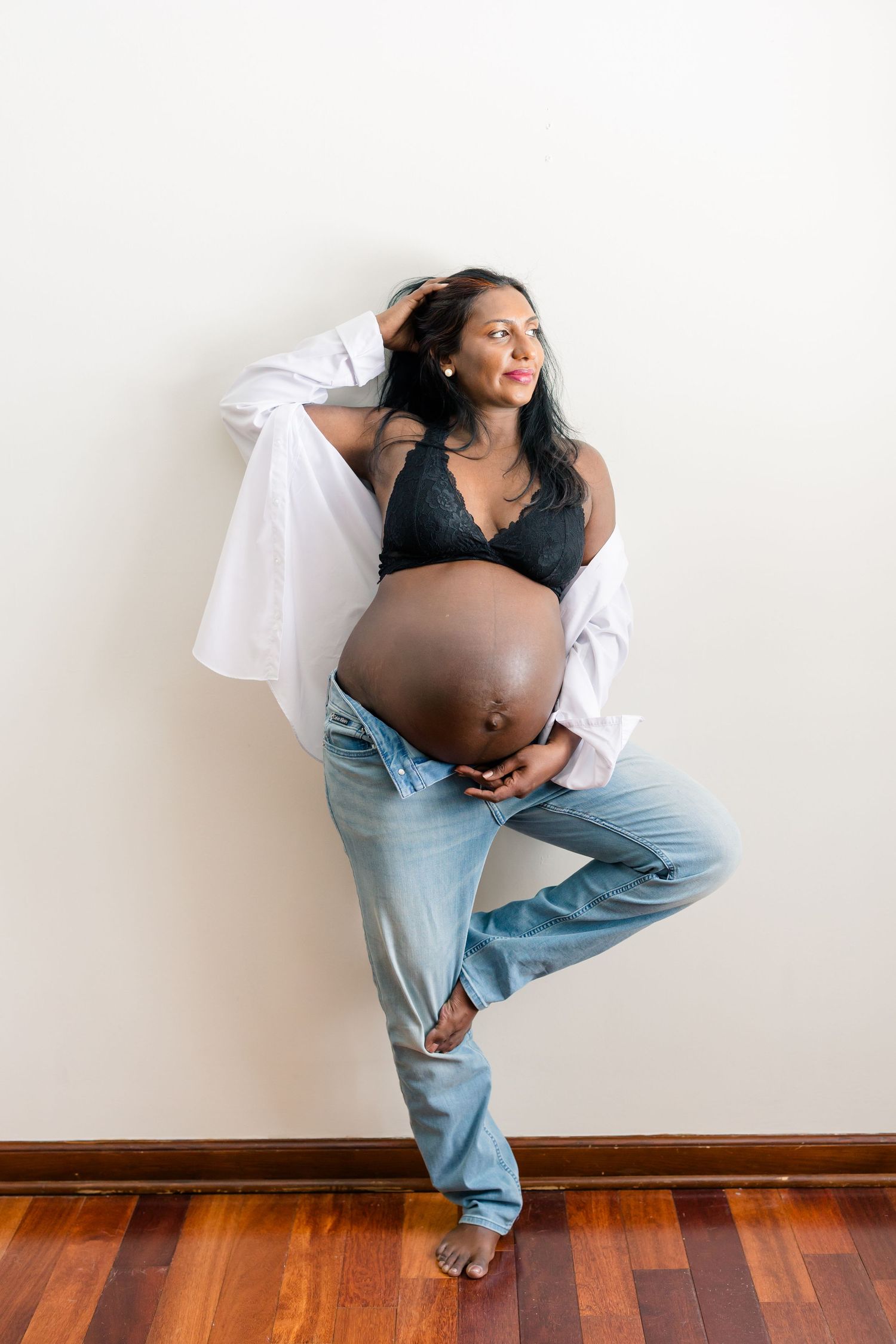 Maternity photo series showing four poses in casual jeans and black top against white wall with hardwood floors.
