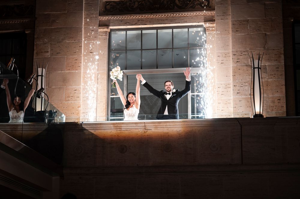 A joyful couple celebrates at their wedding reception with sparklers illuminating the stone building's window behind them.