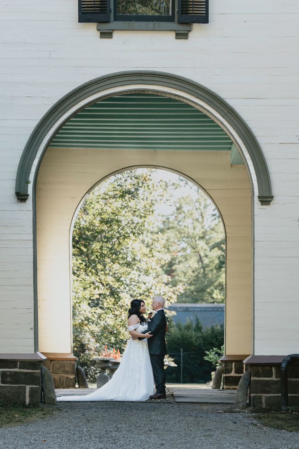 A romantic wedding couple stands in a series of arched white architectural frames surrounded by lush garden scenery.