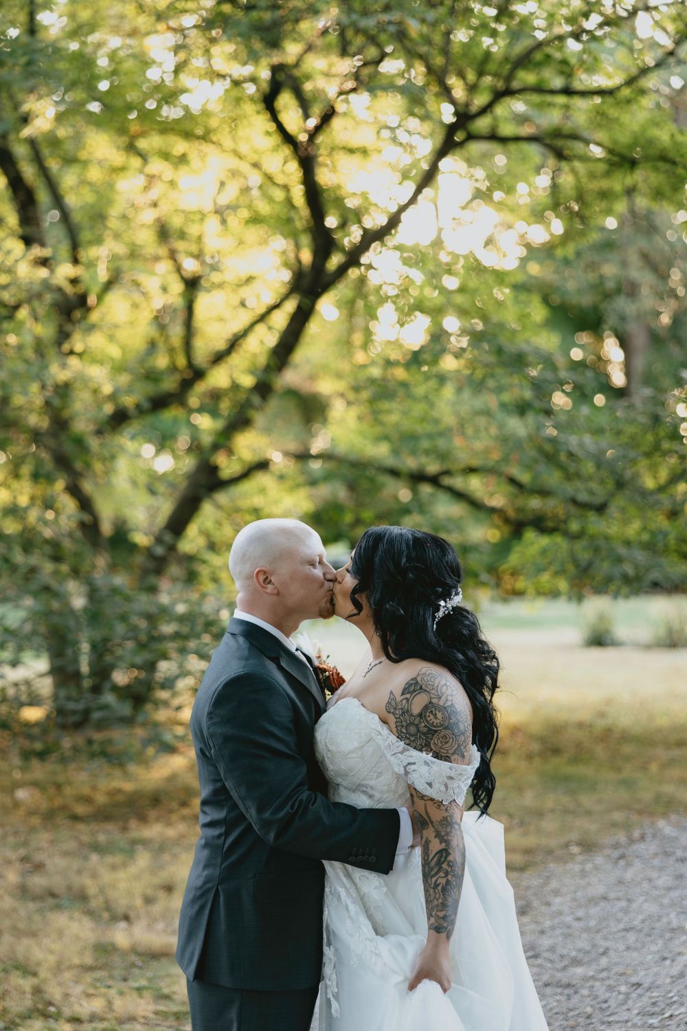 A romantic outdoor wedding moment captured under green trees on a gravel path during golden hour.