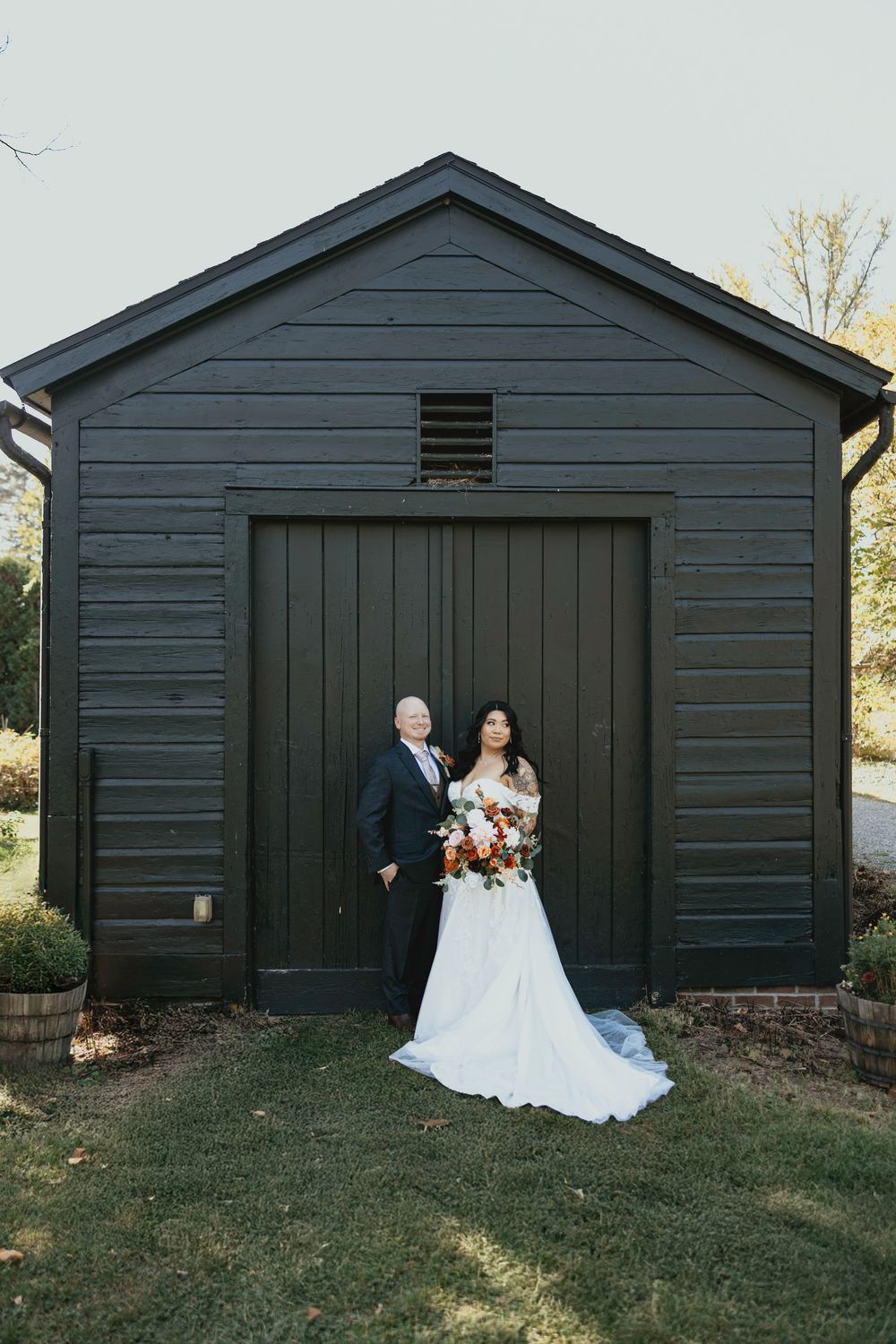 A wedding couple poses for photos in front of a dark gray barn-style building surrounded by grass.