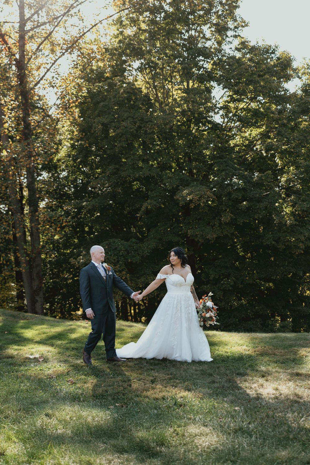 A couple in formal wedding attire walk together through a sunlit grassy hillside with trees in the background.
