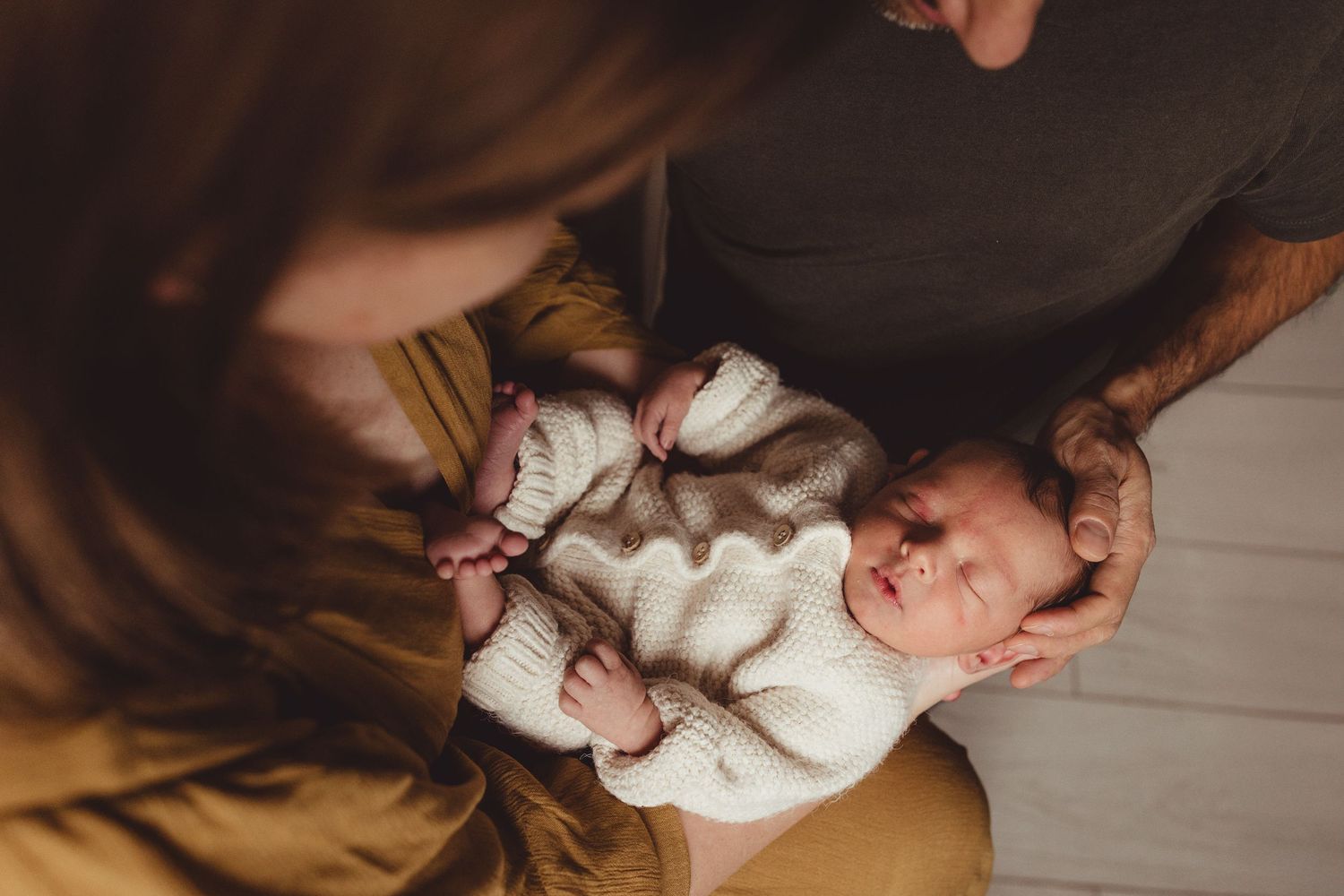 A couple holds their newborn baby in a dimly lit room with natural window light.