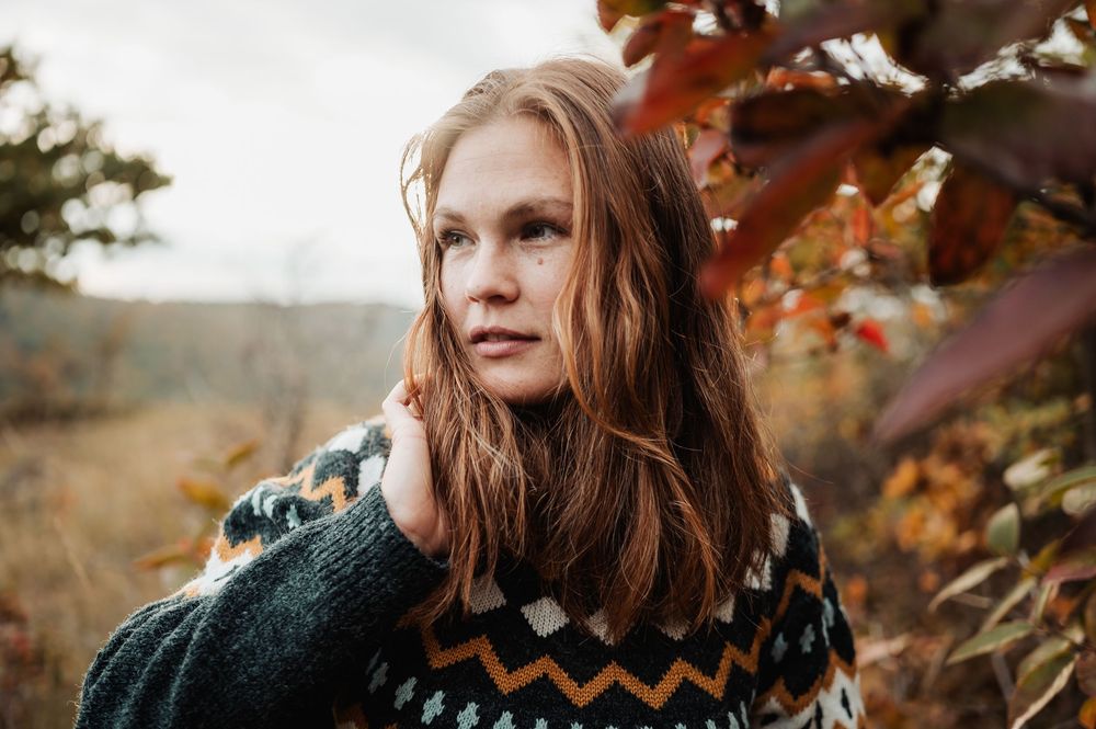 A person in a cozy sweater stands among autumn foliage with golden sunlight filtering through the trees.