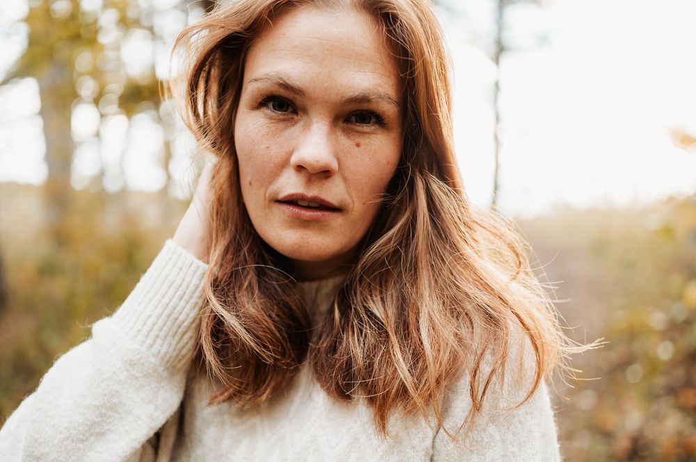 A person in a cozy cream sweater poses outdoors during autumn with soft natural lighting and blurred trees in background.