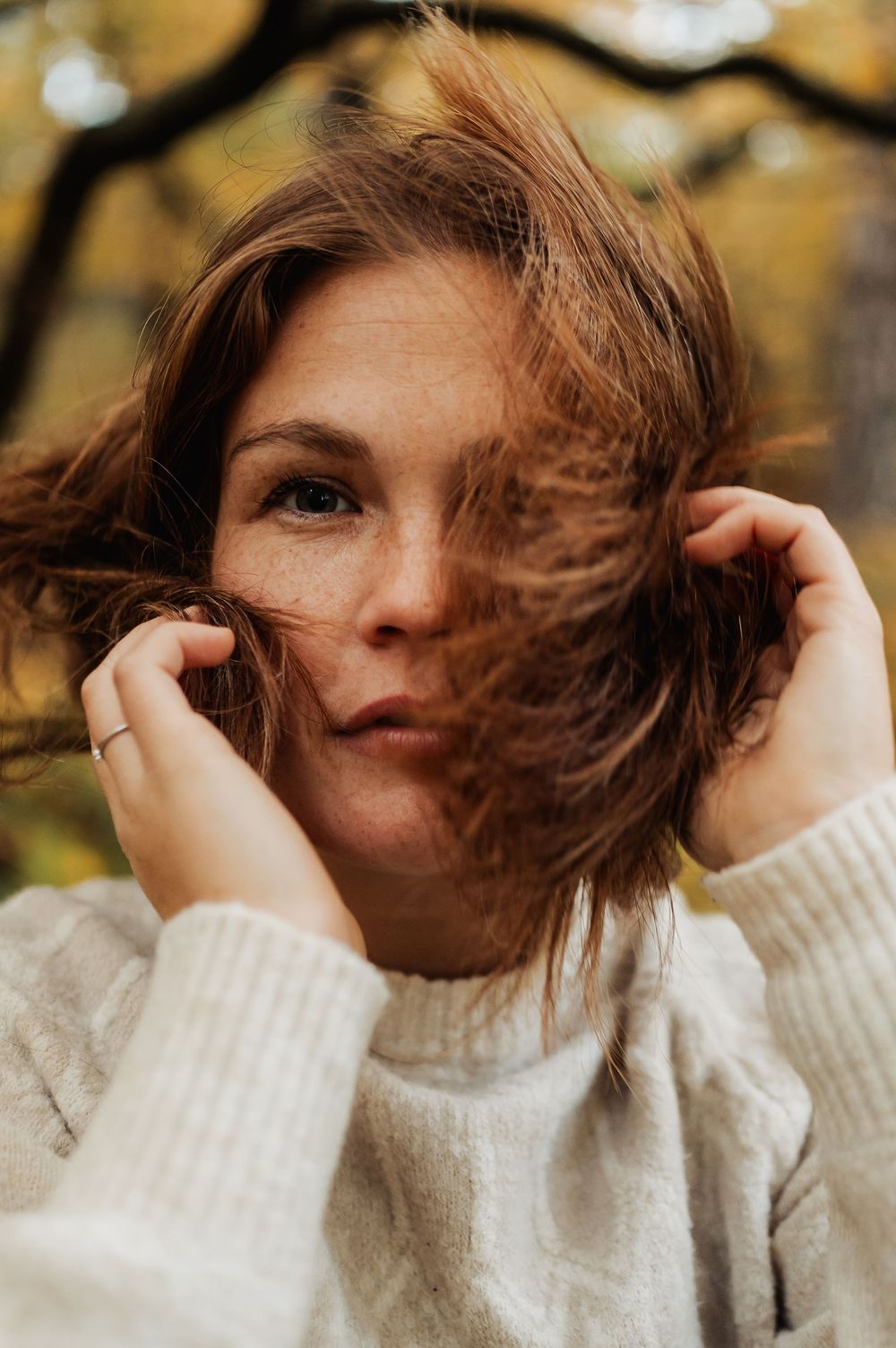 Person in cream sweater with windswept brown hair against autumn background in artistic portrait series.