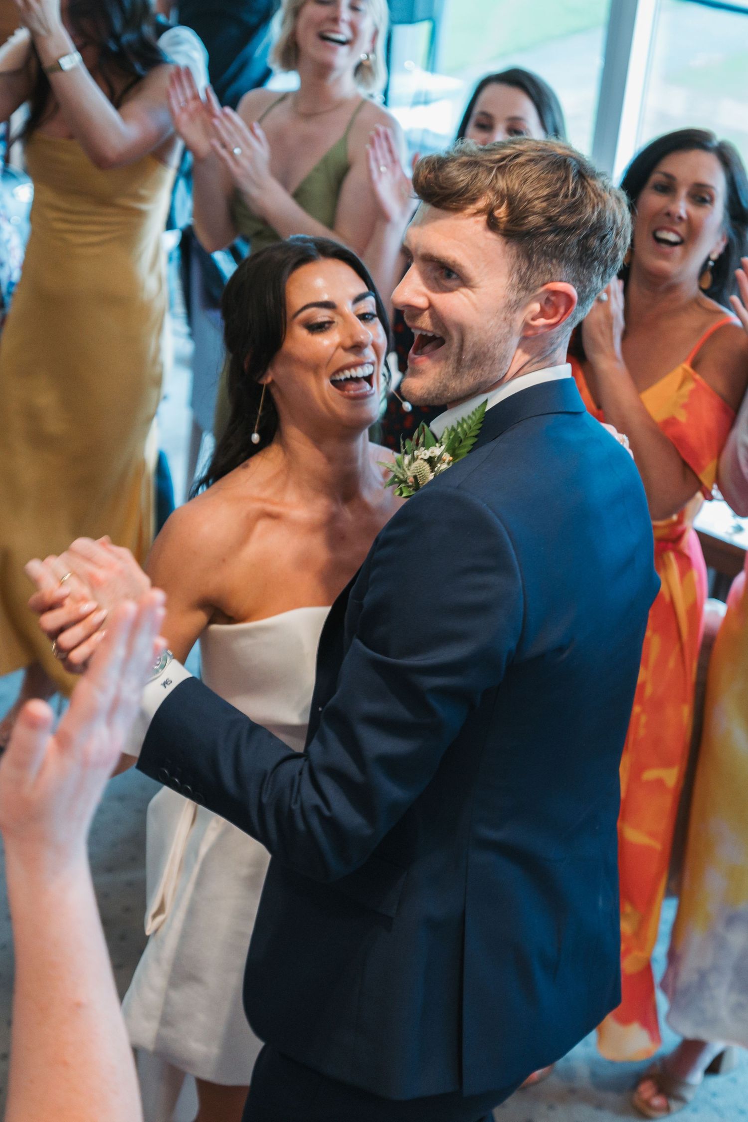Joyful couple dancing and laughing at wedding reception while guests in colorful dresses cheer in background.