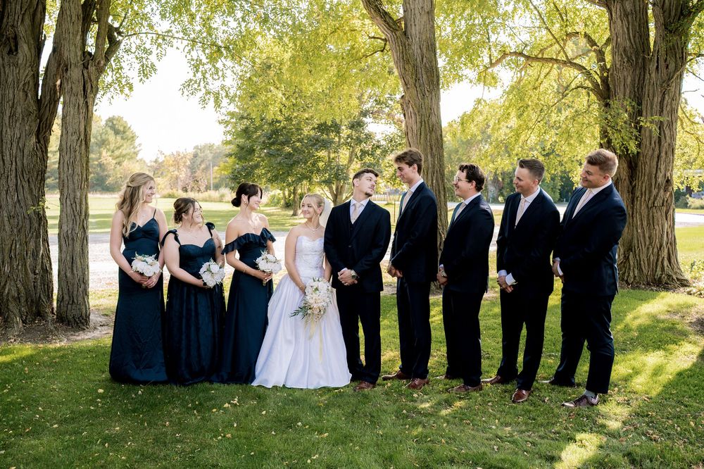 Wedding party in formal attire poses together under tall autumn trees in outdoor setting.