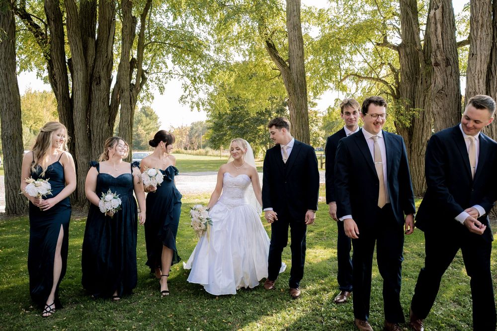 Wedding party in formal attire poses together on grassy lawn with trees and lake in background.