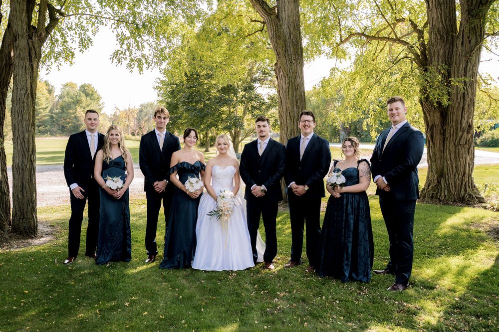 Wedding party poses outdoors in formal attire under large trees during golden hour sunlight.