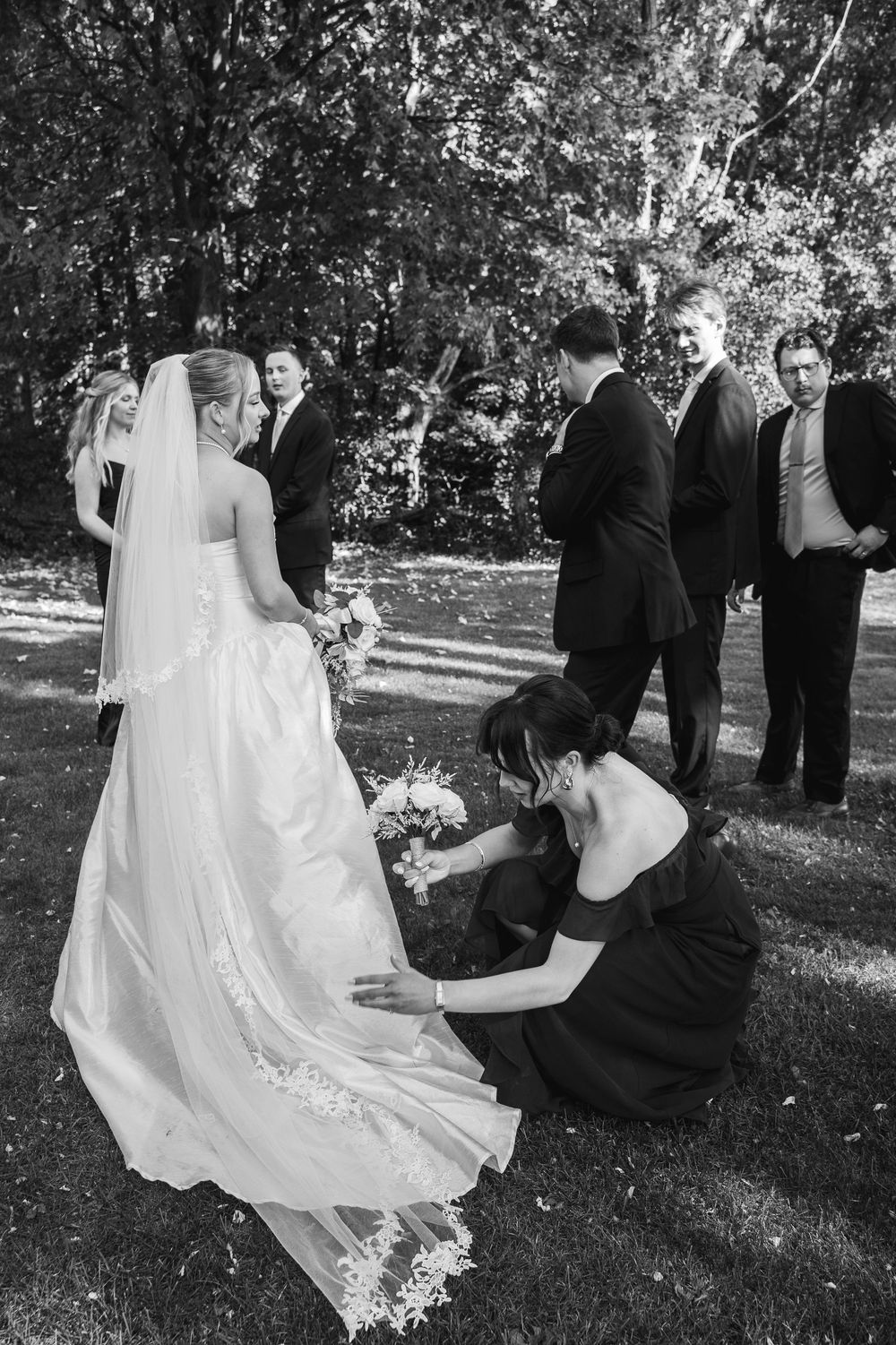 Black and white sequence shows wedding party arranging bride's dress on grass during outdoor photo session.