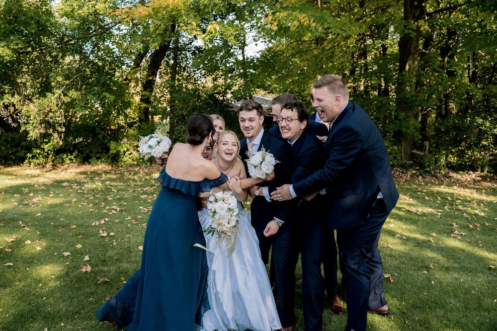 Wedding party poses together outdoors in a natural setting with green trees and sunlight filtering through.