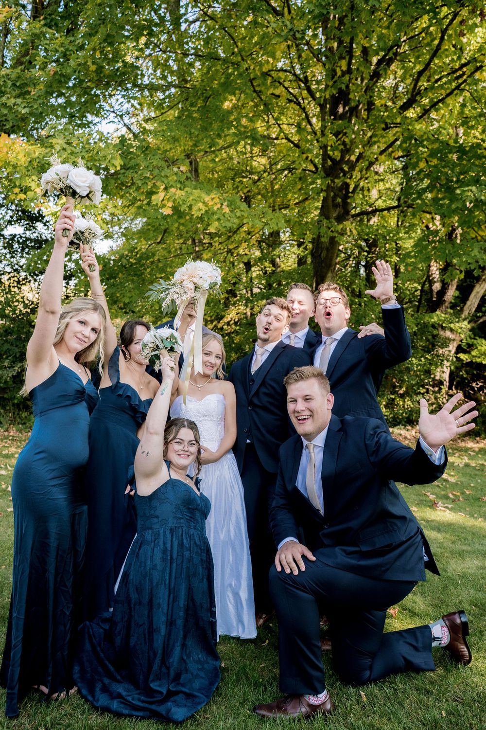 Wedding party poses joyfully on a grassy hill with trees in the background during an outdoor celebration.