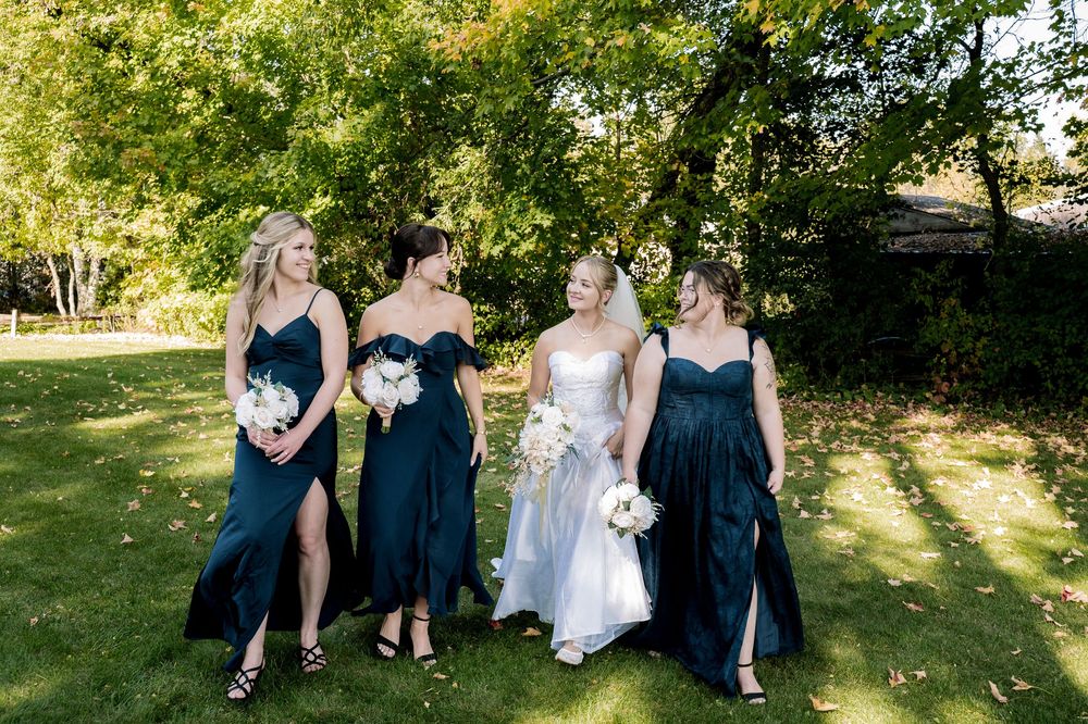 Wedding party members in navy blue dresses and suits pose together on a sunny lawn surrounded by trees.