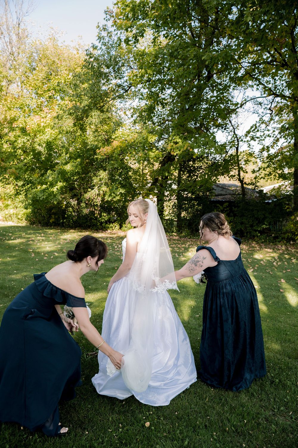 A wedding party sits in a grassy field with the bride in white and attendants in navy blue dresses arranged in a row.