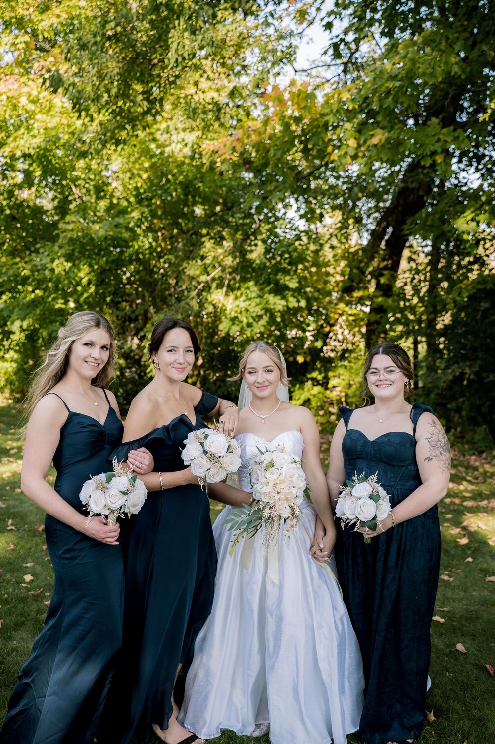 Bridal party stands together under green trees wearing black dresses and white gowns.
