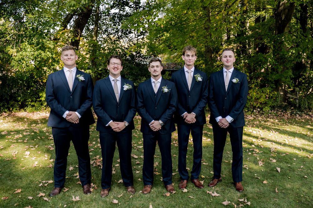 Groomsmen in dark suits and blue ties stand in a line outdoors with green foliage in the background.