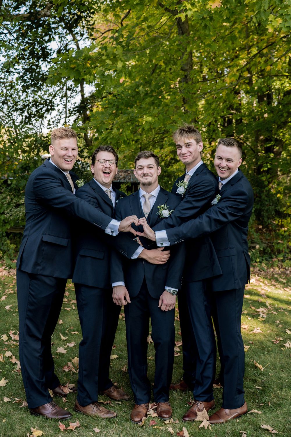 A group in black suits and brown shoes poses playfully together on grass under autumn trees.
