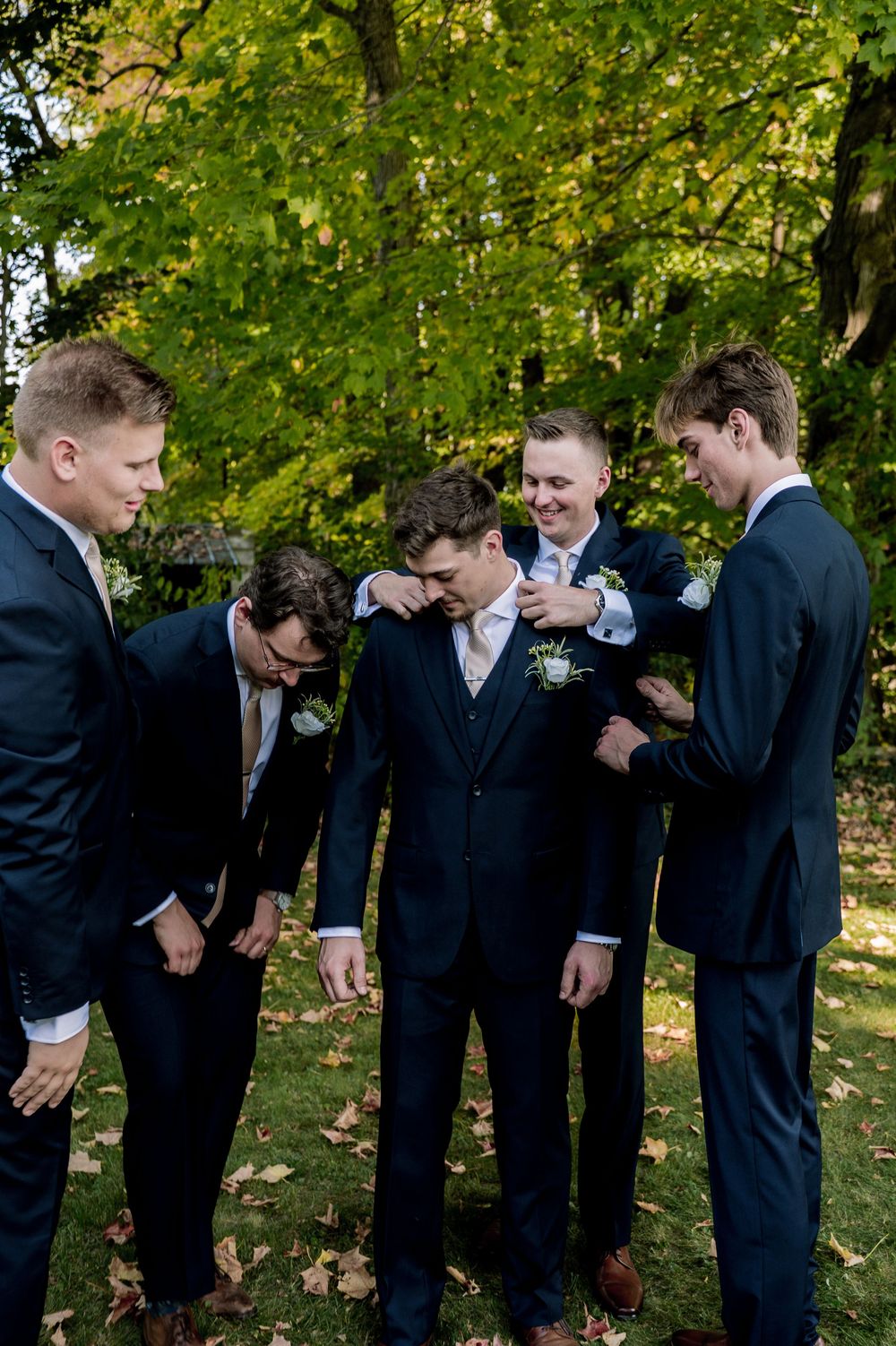 Groomsmen in dark suits lean forward in a playful pose sequence under green trees during a wedding photoshoot.