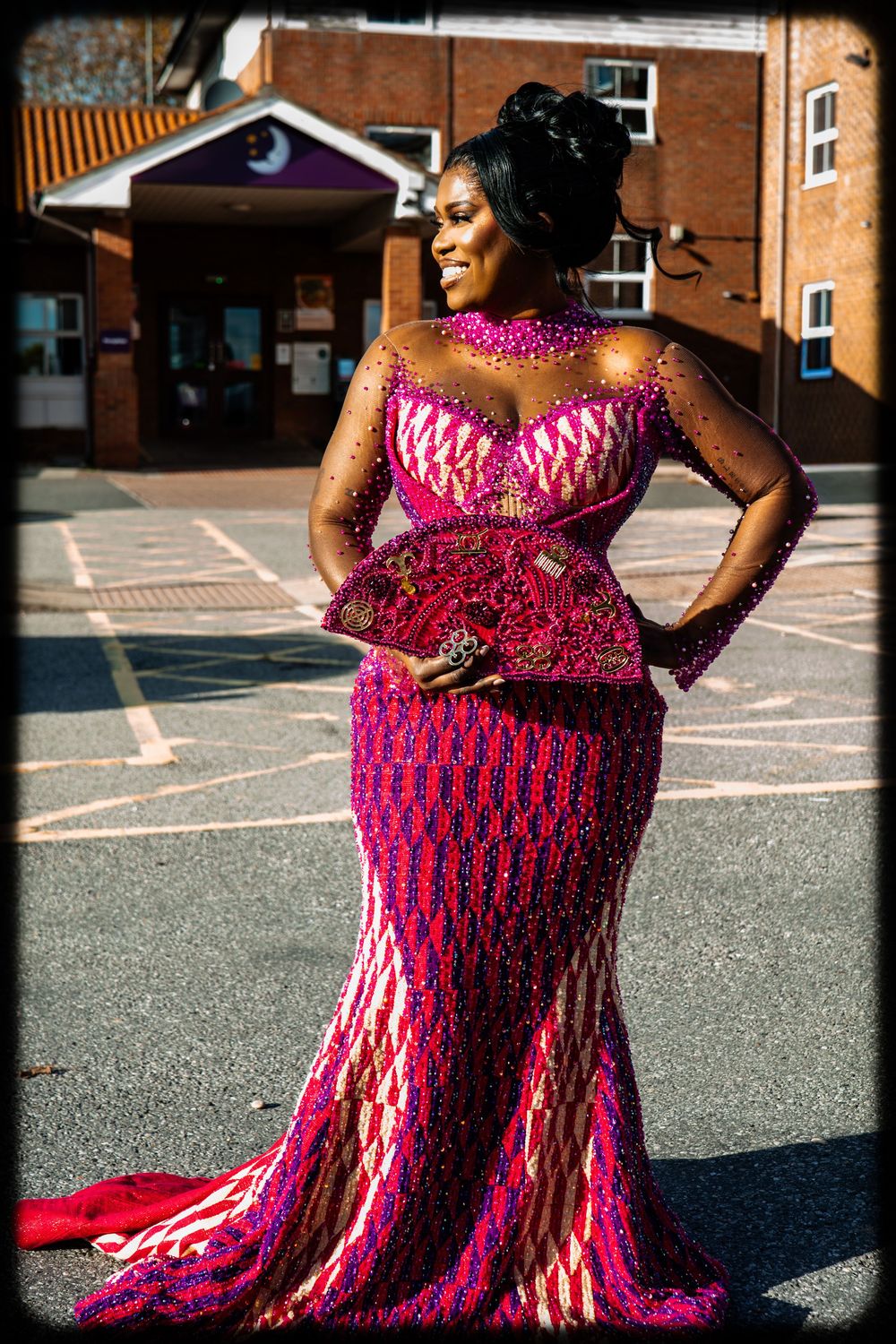 A person in a vibrant pink patterned dress poses outdoors in a parking lot on a sunny day.