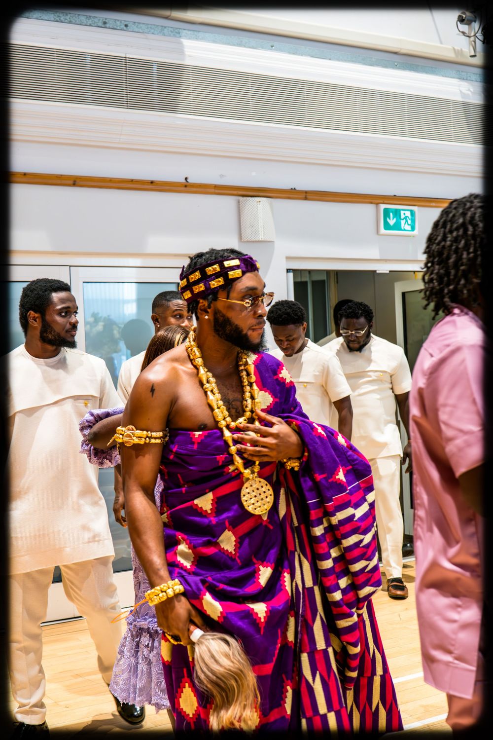 People in traditional African attire with vibrant purple and pink patterns gather at an indoor event.