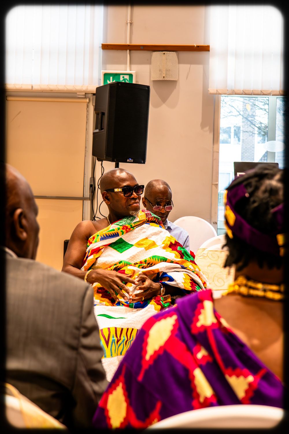 People gathered at an event wearing vibrant traditional African kente cloth and colorful attire in an indoor setting.