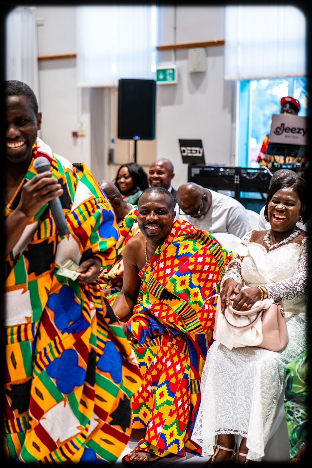 A group of people wearing vibrant kente cloth celebrate joyfully together at an indoor gathering.