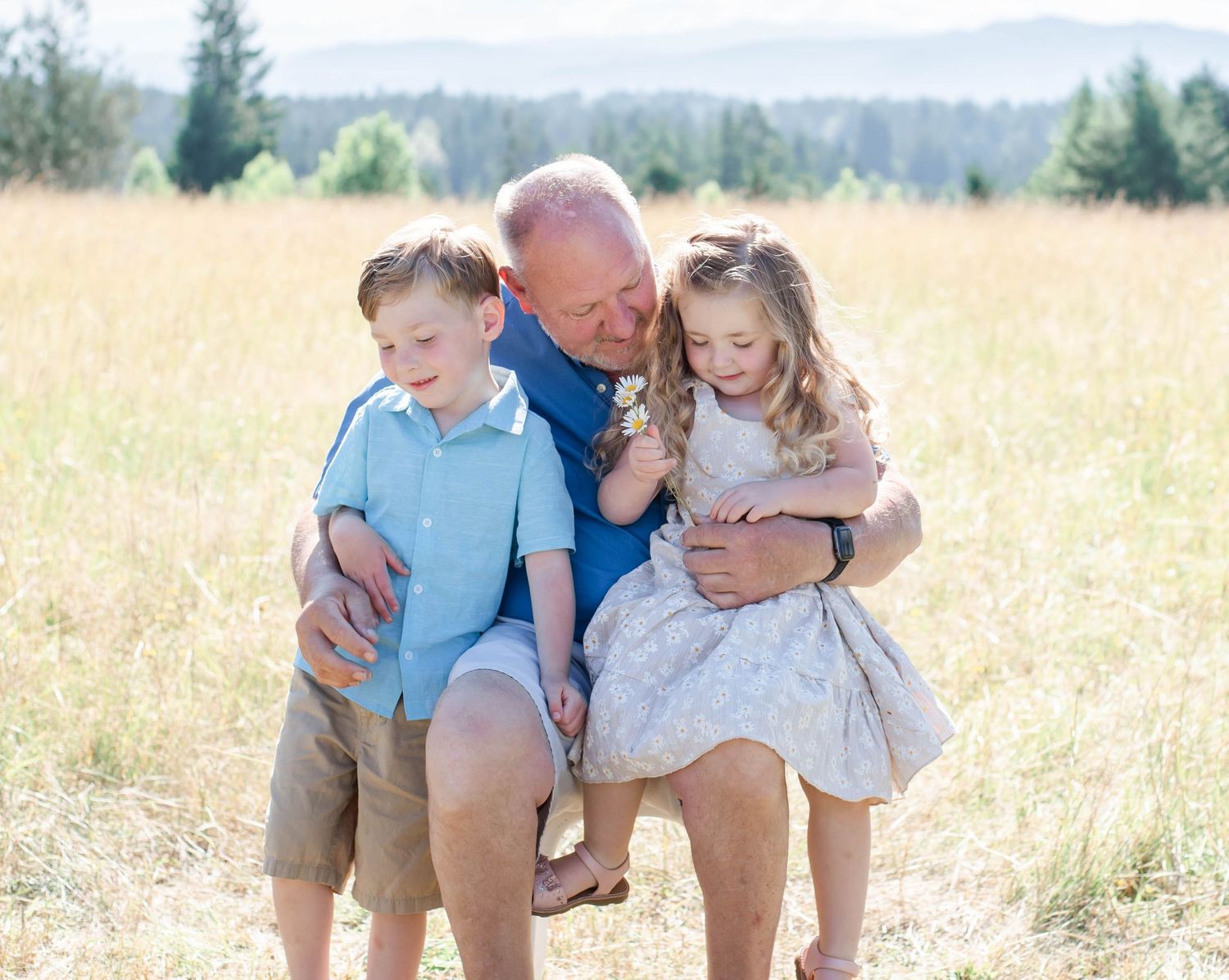 Family sitting together on blanket in sunny golden field with mountains and evergreen trees in background.