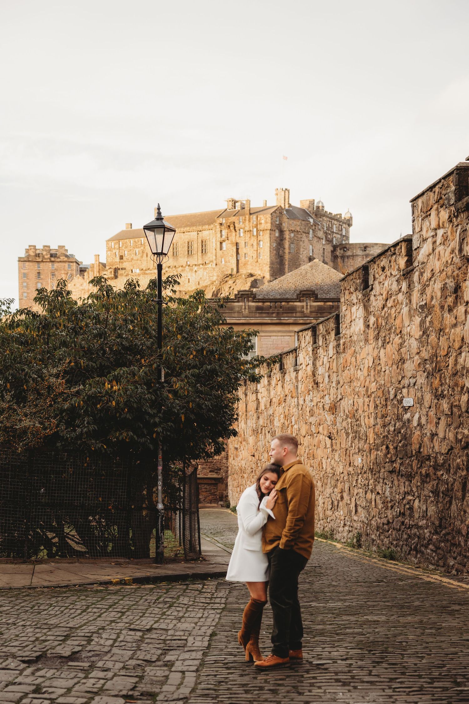 A couple embraces on a cobblestone street near historic stone walls and castle buildings in Edinburgh, Scotland.