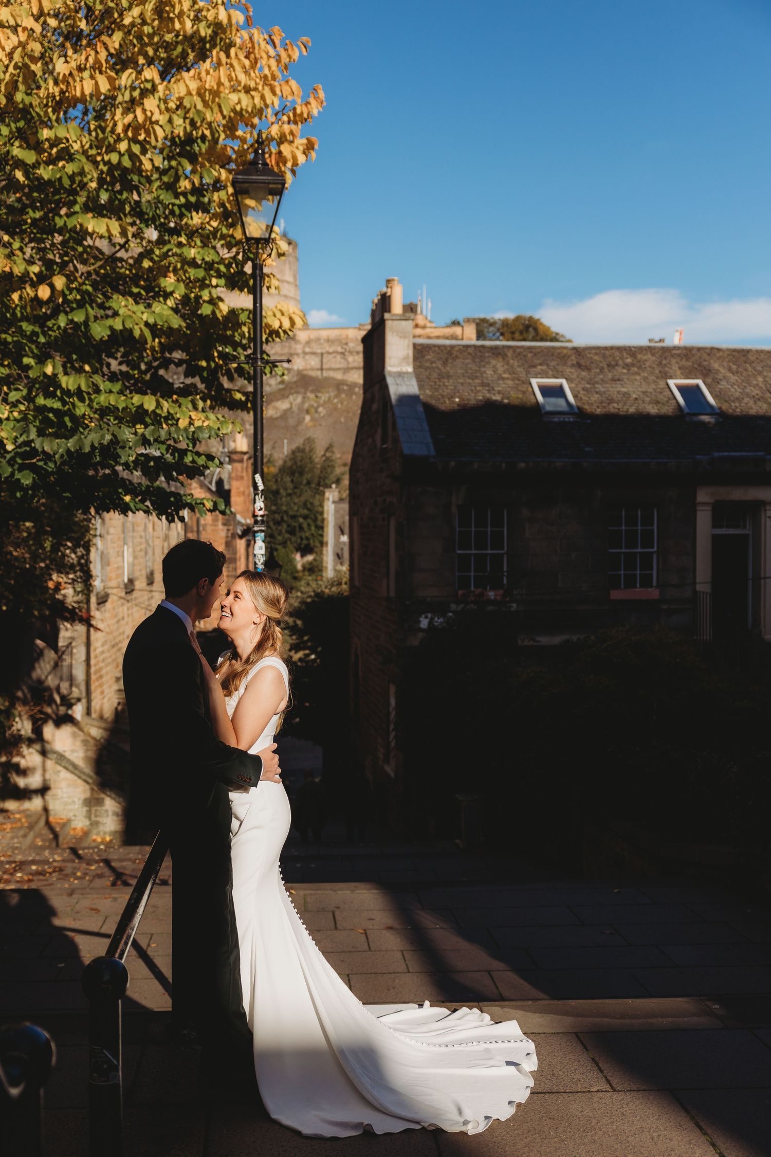 A wedding couple shares an intimate moment at sunset against a stone building with autumn foliage overhead.
