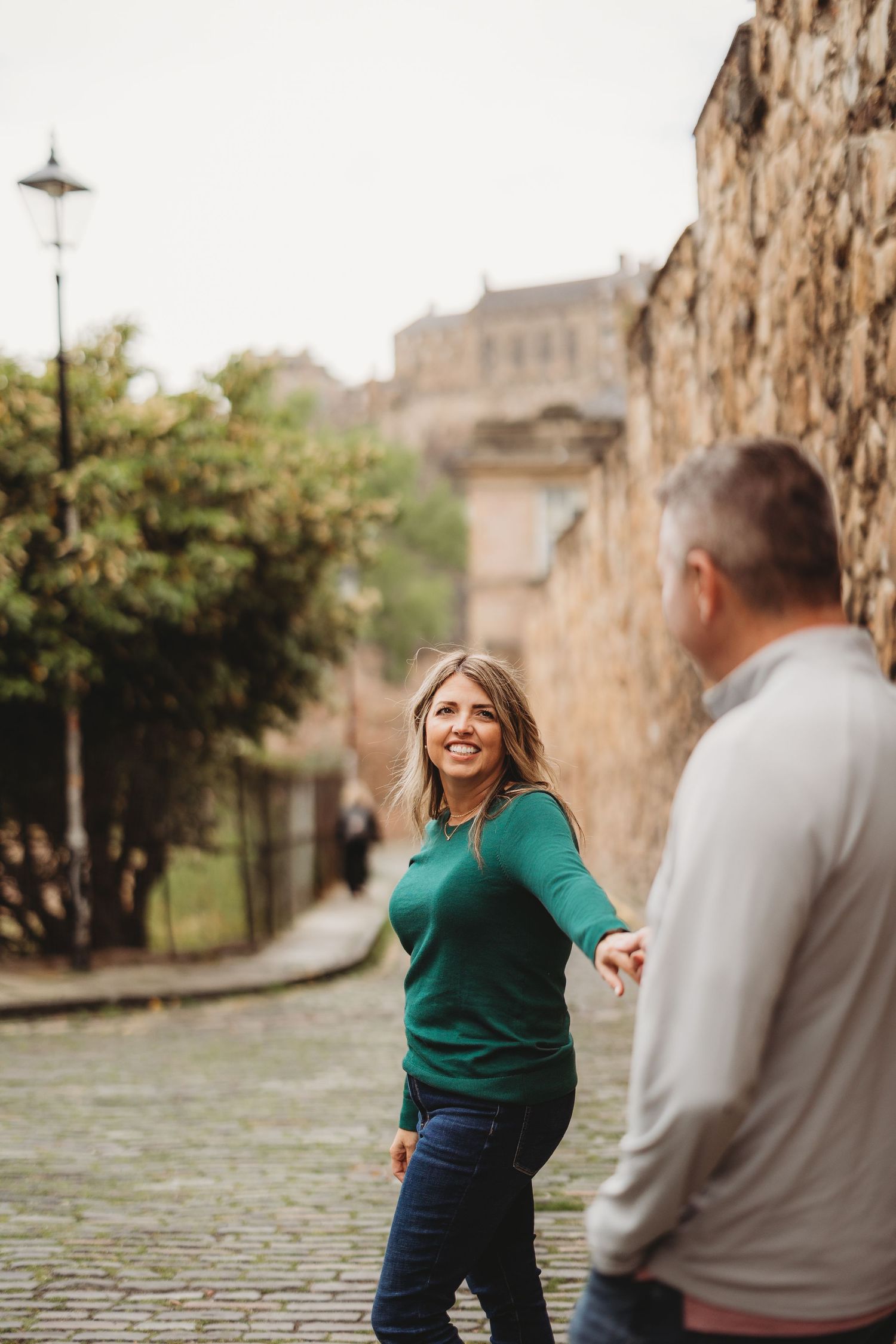 Two people enjoying a casual stroll along a cobblestone street with historic stone walls and architecture in the background.