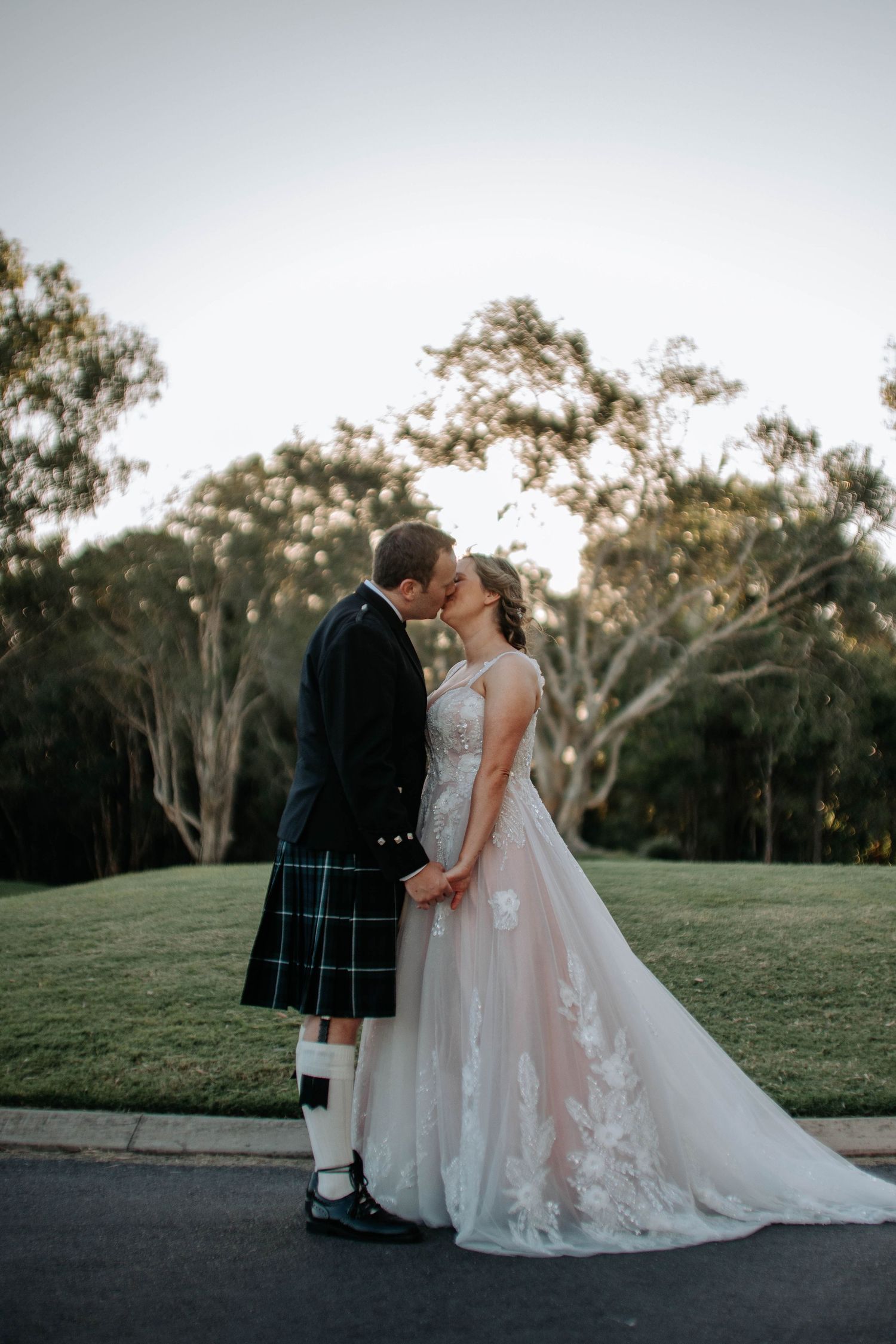 A couple in traditional wedding attire share an intimate moment on a grassy hillside at sunset.