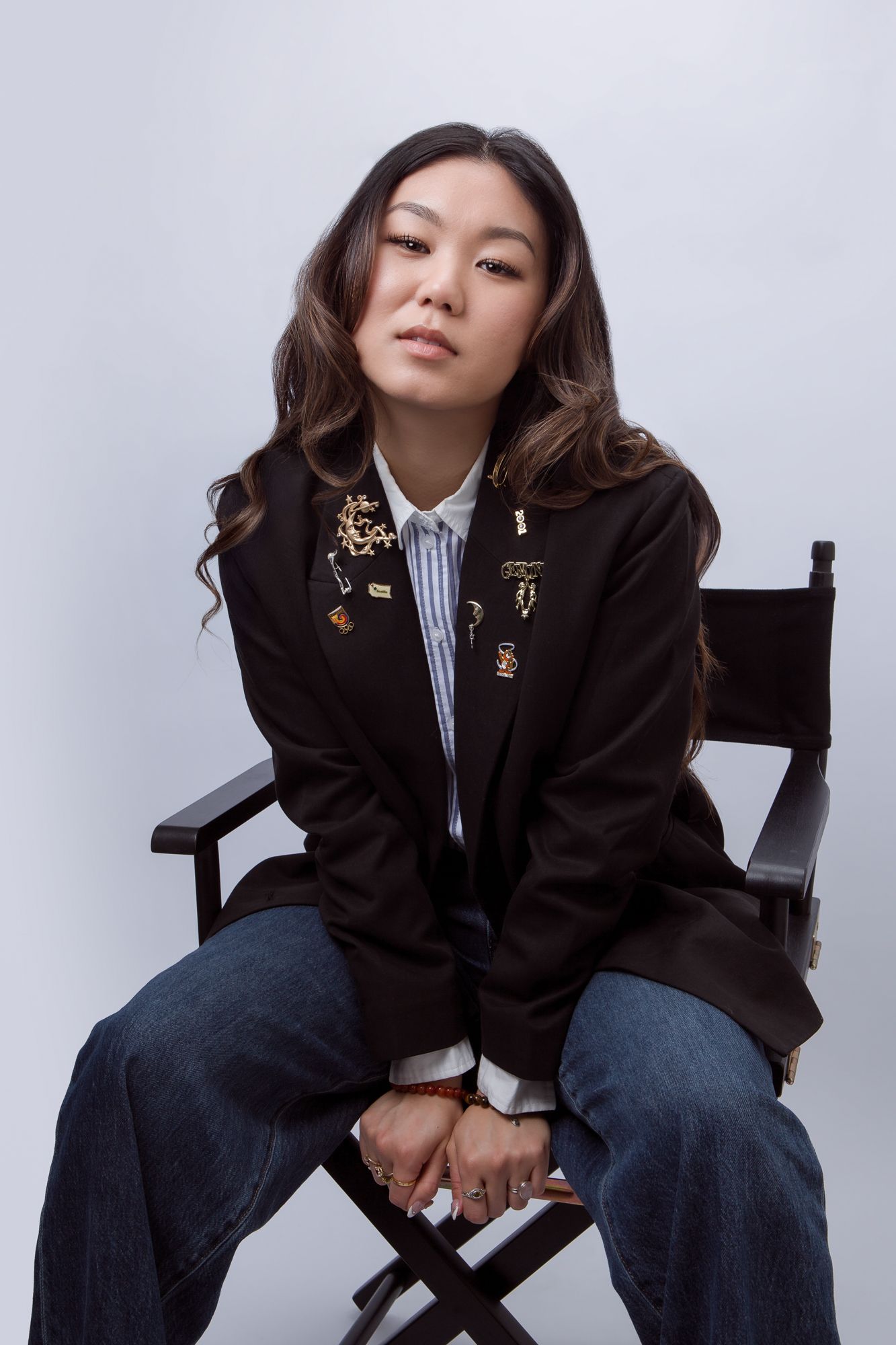 A person in a black blazer and striped shirt poses casually on a chair against a white studio backdrop.