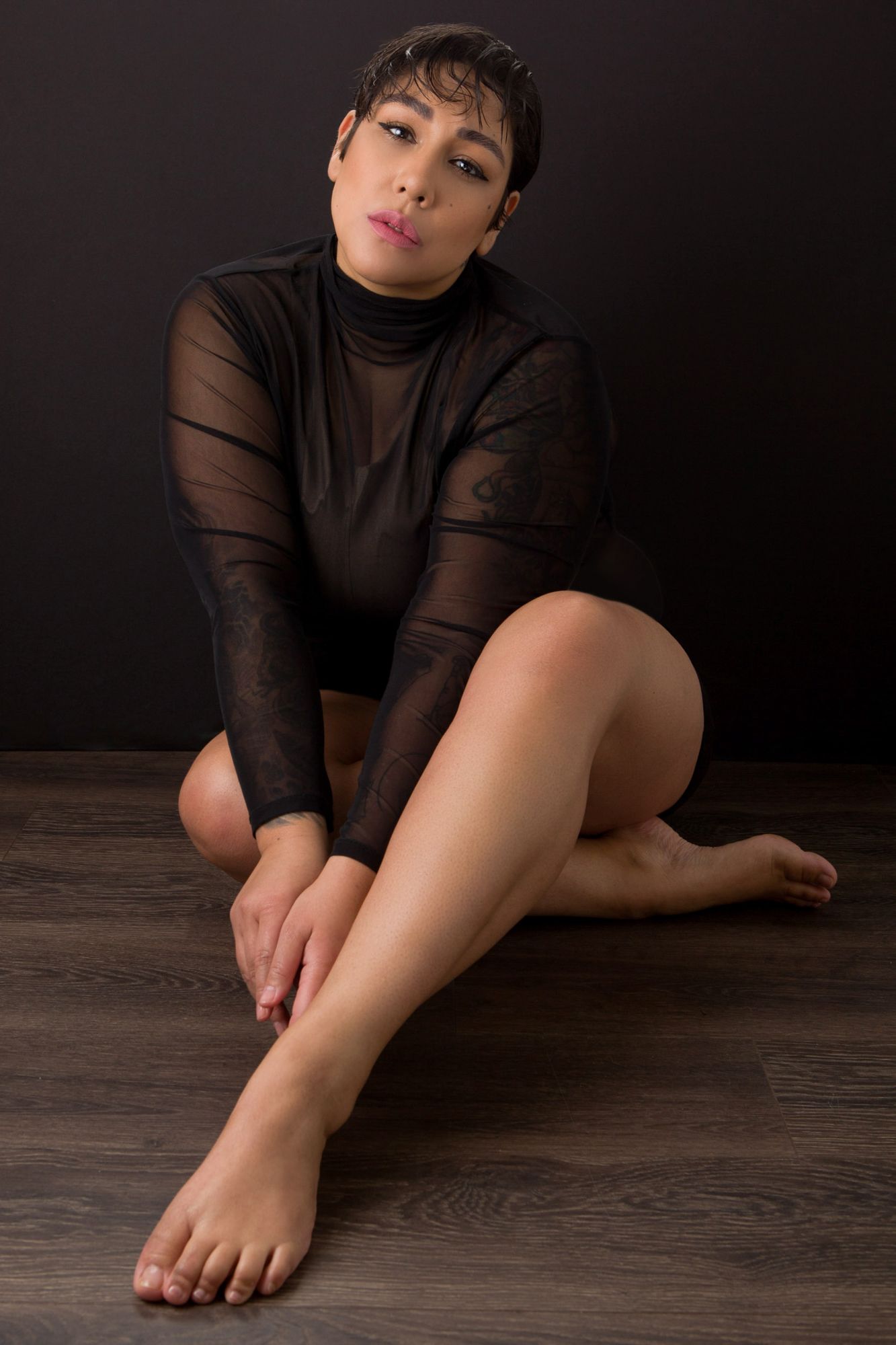 A person in a black long-sleeved top sits on a dark wooden floor while showing their bare feet.