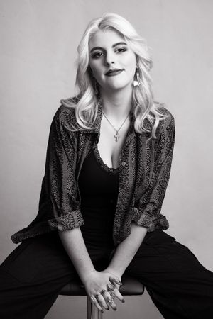 Black and white portrait of a person with platinum blonde hair wearing a patterned jacket and black top against a plain backdrop.