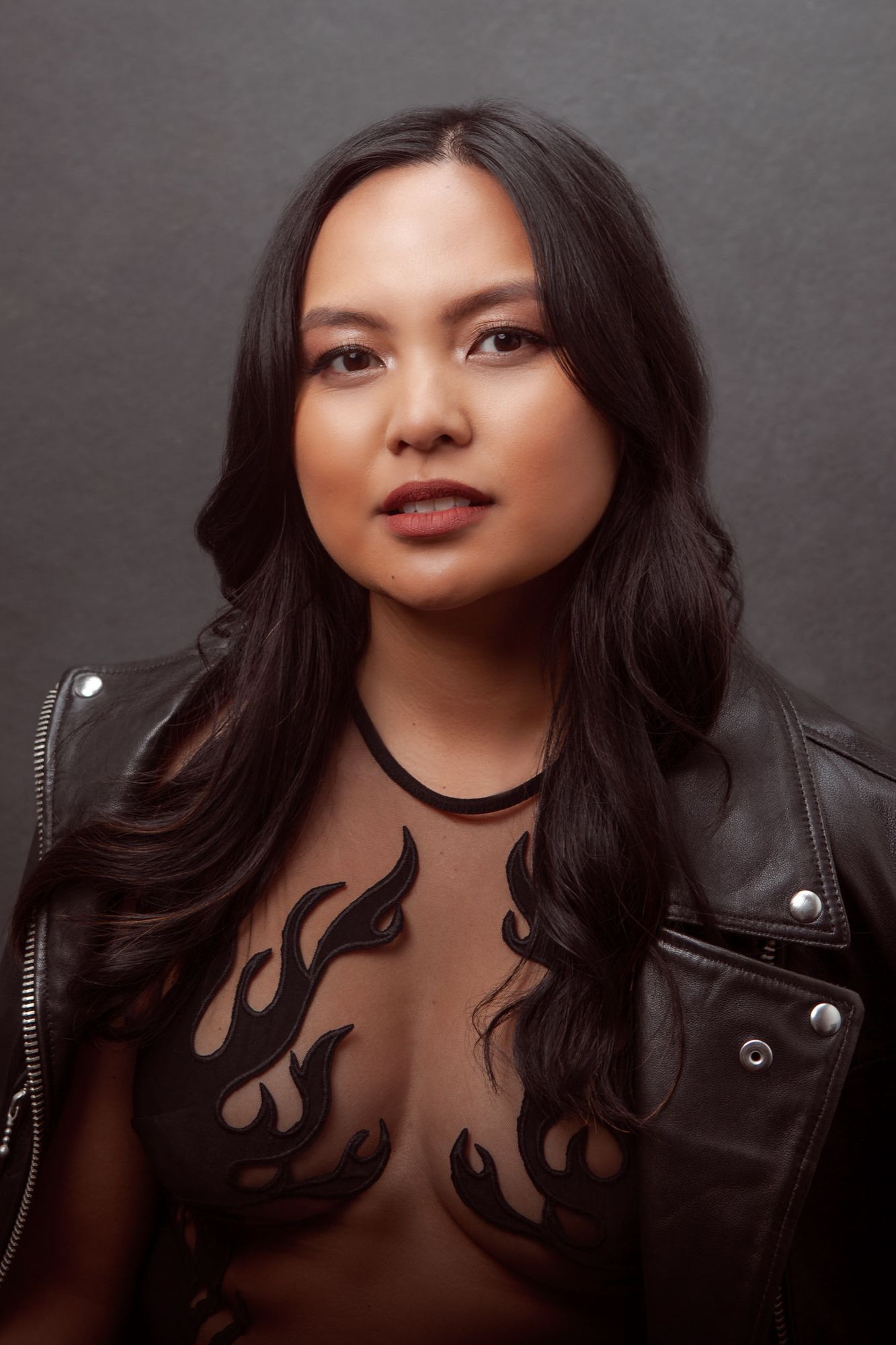 Portrait of someone in a black leather jacket and sheer top with flame designs against a dark gray background.