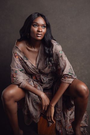 Artistic portrait of someone wearing a floral wrap dress seated on a wooden stool against a dark backdrop.