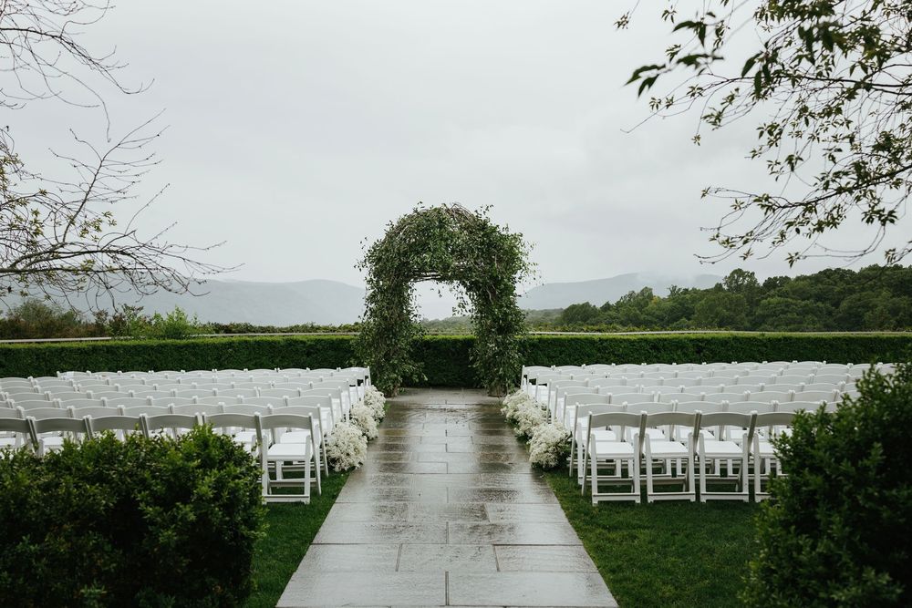 White chairs line both sides of a wet walkway leading to a lakeside wedding ceremony setup in rainy weather.