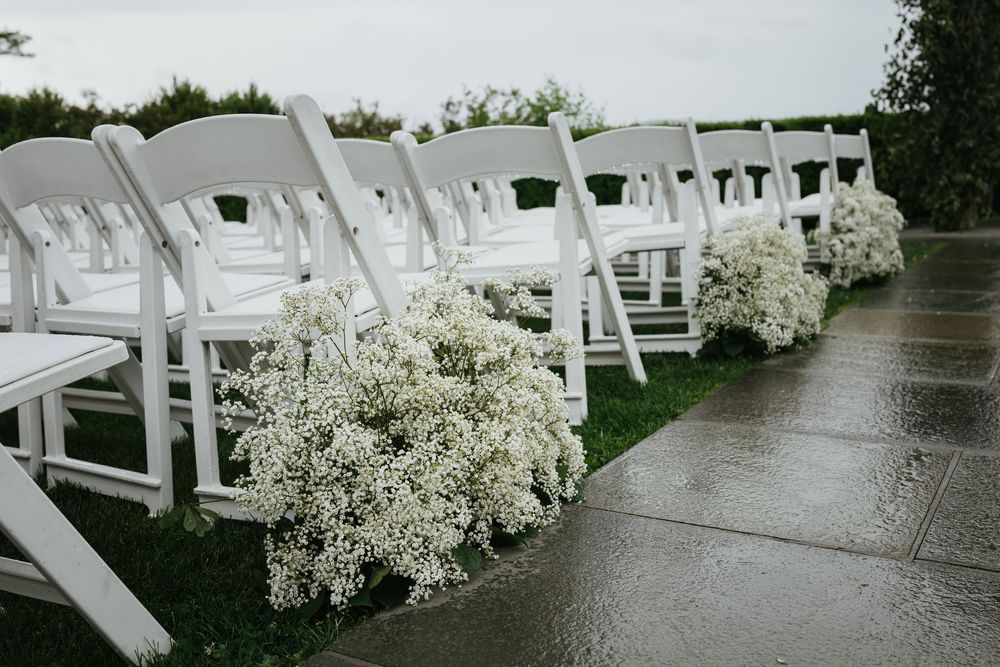 White wedding ceremony chairs line an outdoor aisle decorated with baby's breath floral arrangements on a rainy day.