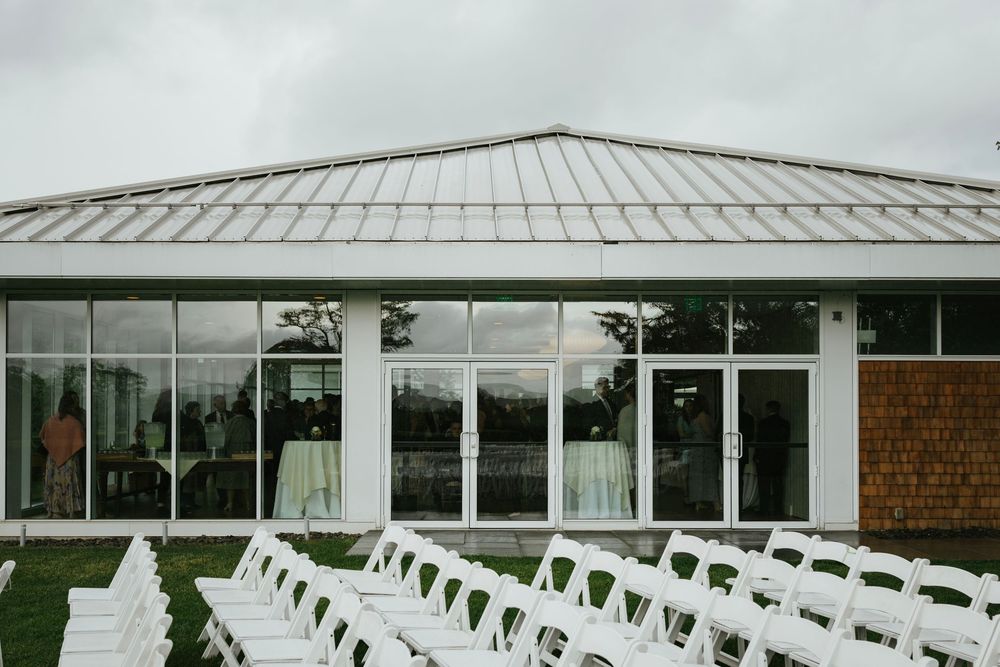 White chairs arranged in rows facing a modern glass-walled event venue with a hip roof and brick accent wall.