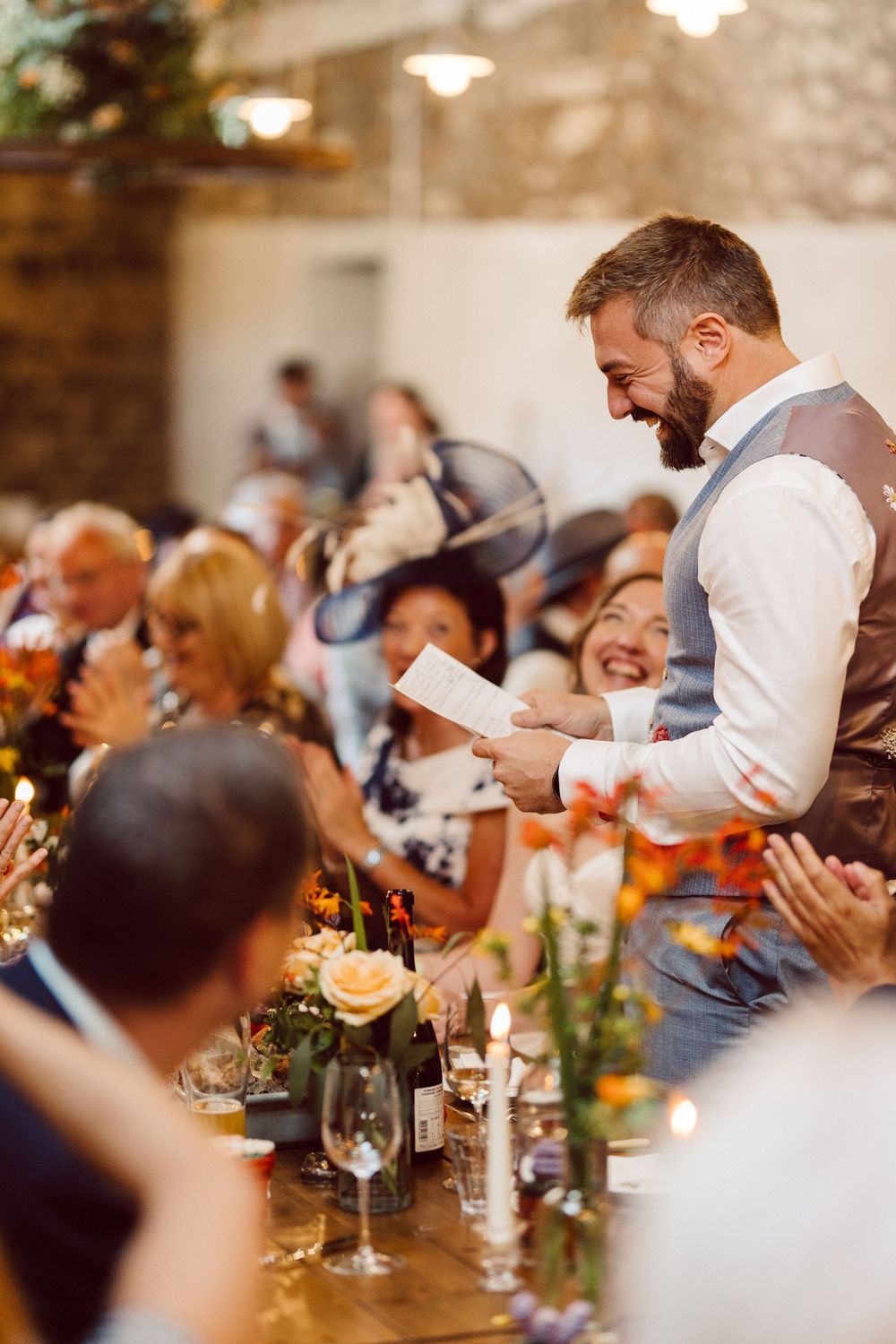 Wedding reception guests seated at long tables enjoy dinner and conversation in a rustic venue with candlelight.