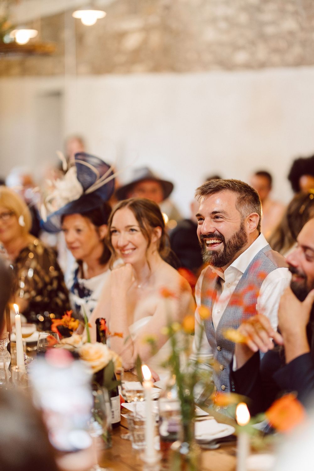 Wedding guests laugh and smile during dinner at a rustic stone-walled reception venue.