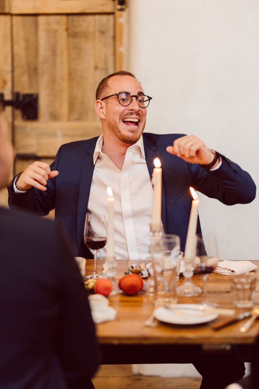 A guest in a navy blazer gestures animatedly while seated at a wedding reception dinner table.