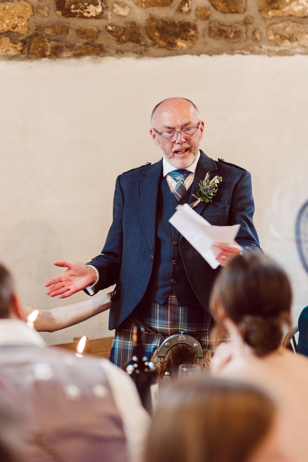 A speaker in a navy suit and kilt delivers an animated speech at a wedding reception.