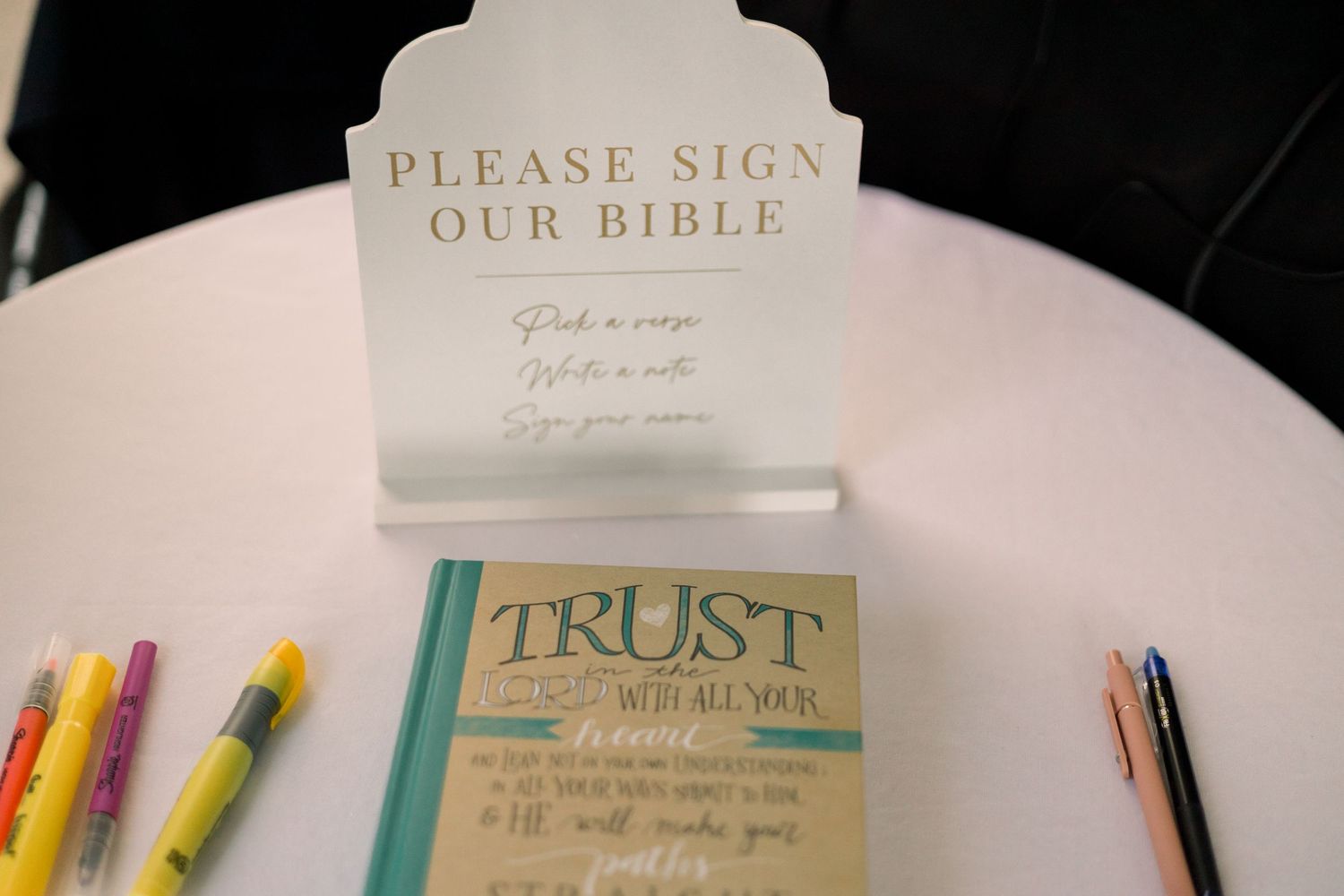 Wedding guest book and colorful markers displayed on table with inspirational quote decor and Bible signing station.