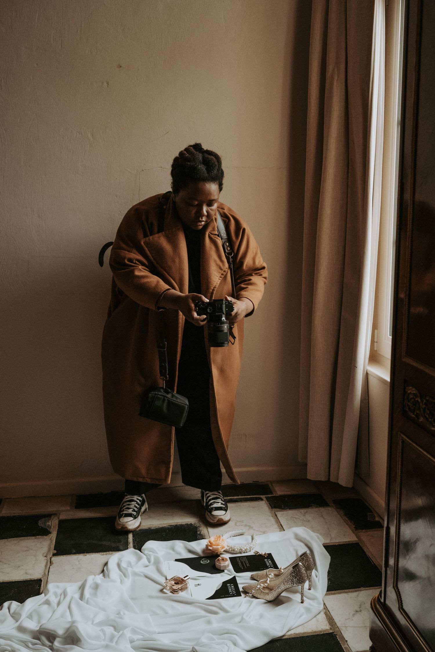 A photographer in a brown jacket takes pictures of items laid out on a white sheet near a window.
