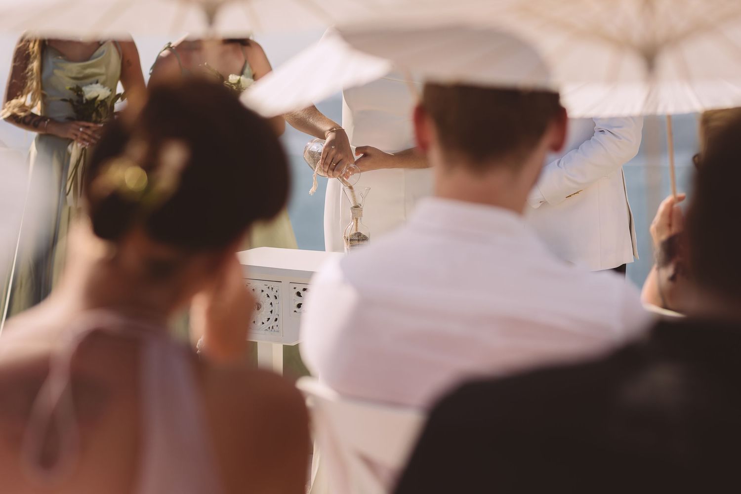 A view of a couple pouring sand into a jar at a destination wedding in Santorini viewed through a gap between guests.