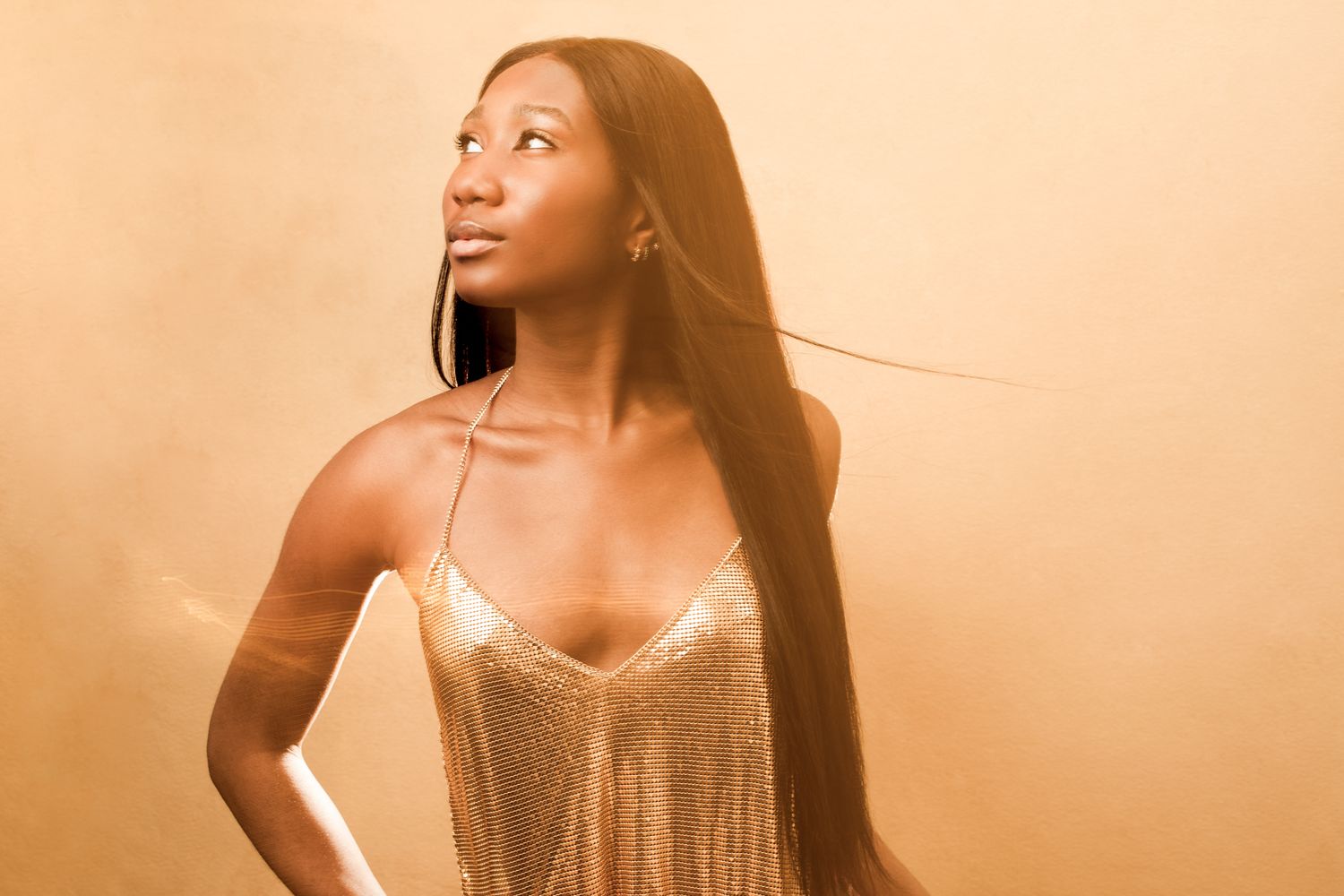 A woman in a sparkly gold dress poses against a warm sepia-toned background with dramatic lighting.