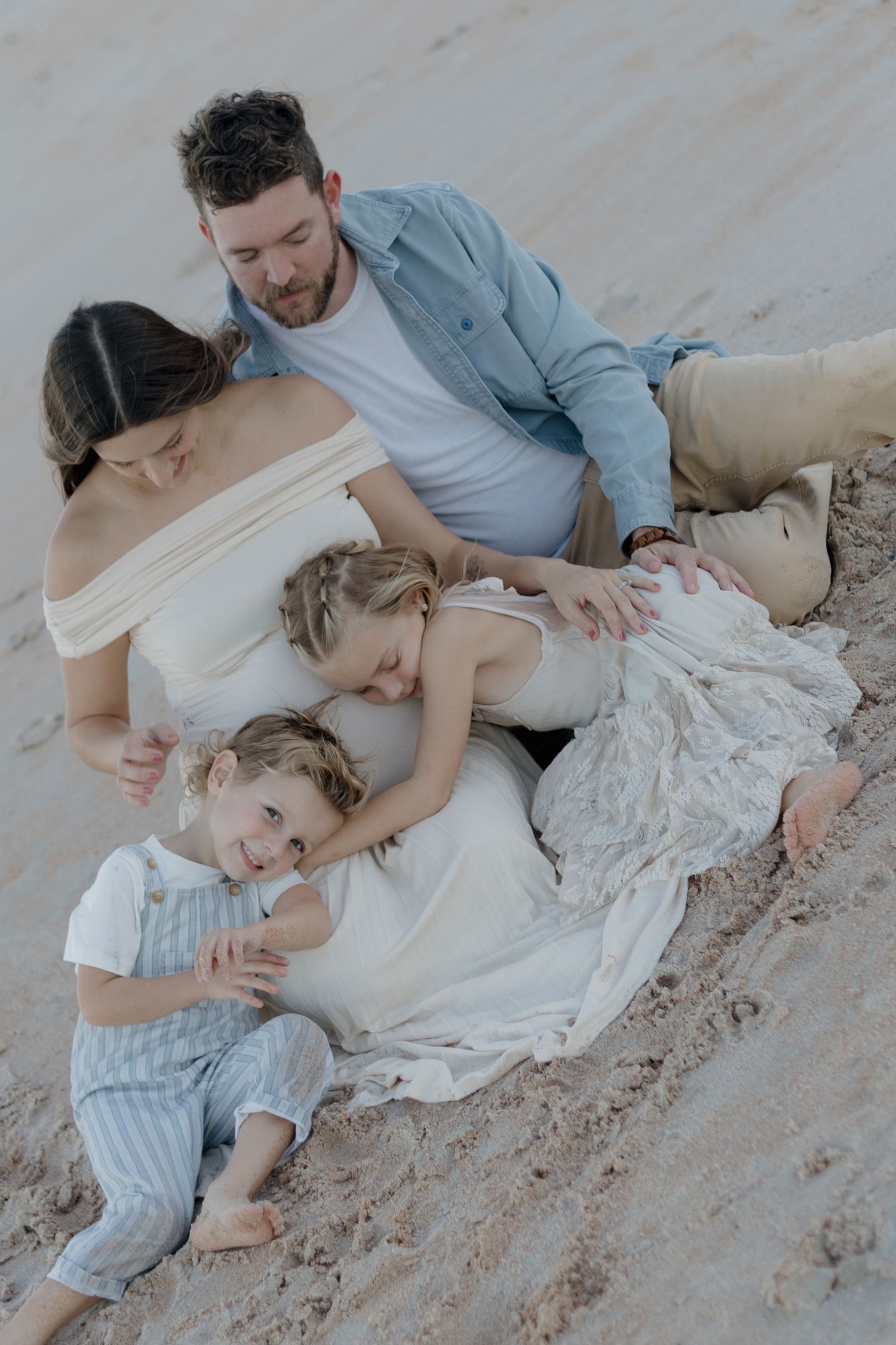 A tender family moment captured on a sandy beach with soft natural lighting and flowing white fabric.